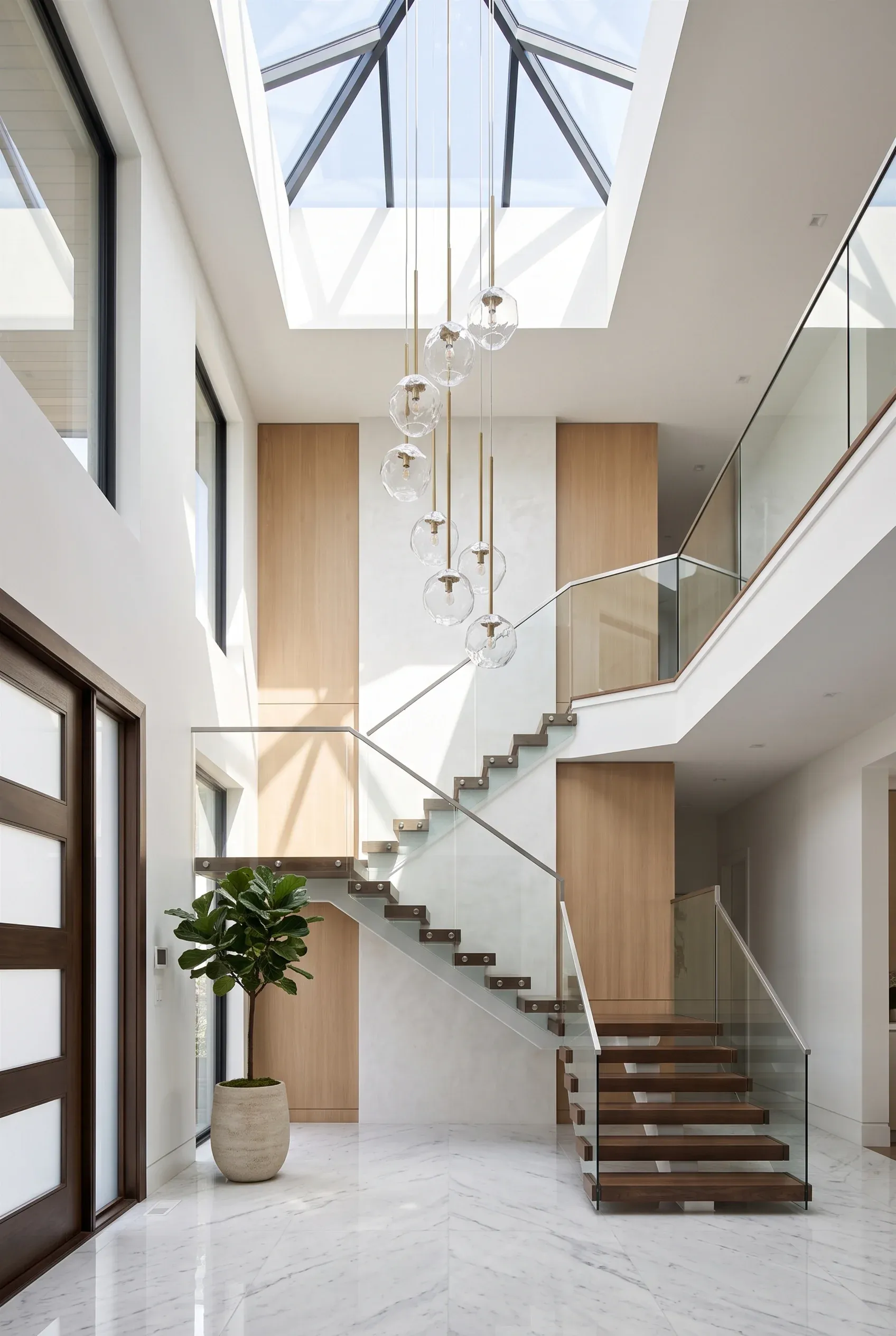Vestíbulo moderno de doble altura con escaleras flotantes de madera, barandales de vidrio, tragaluz, iluminación colgante y piso de mármol, diseño de RBA Residences en Zapopan.