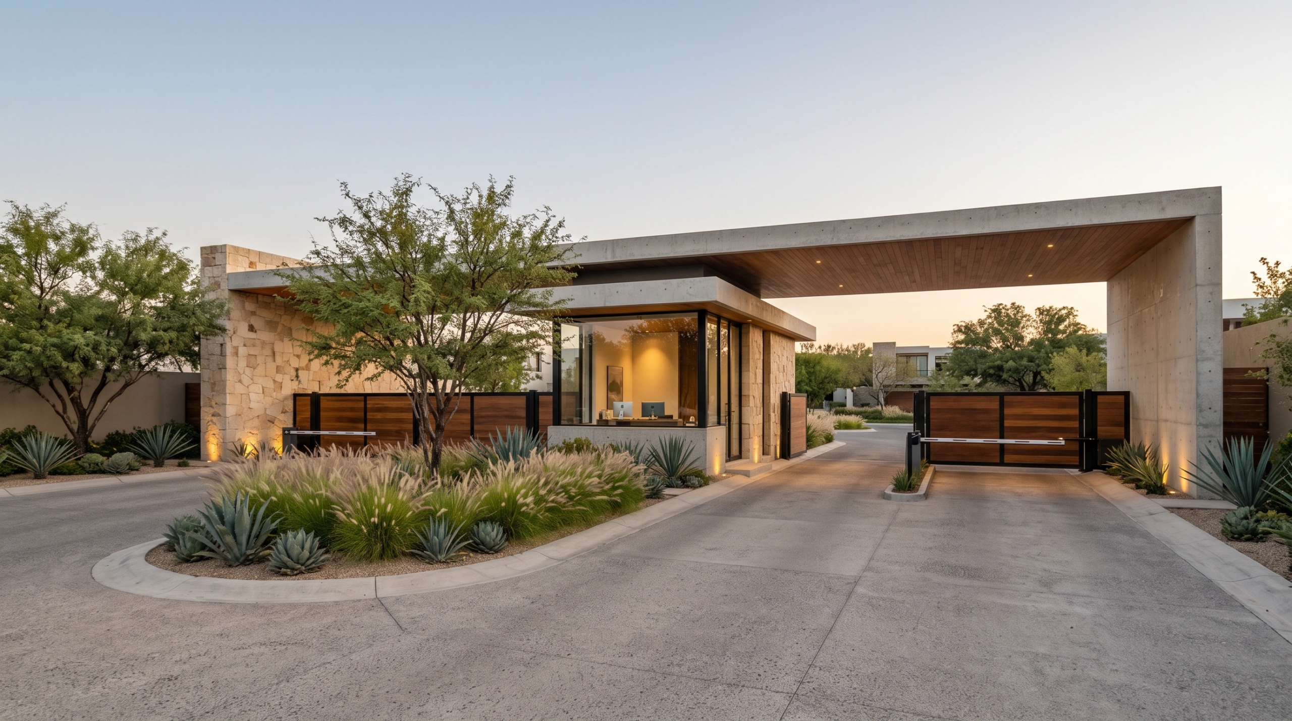 Vista de entrada arquitect&oacute;nica moderna para una comunidad residencial privada con voladizo de concreto, muros de piedra, caseta de vigilancia de vidrio central, portones de seguridad de madera y agaves, acceso exclusivo de RBA Residences en Zapopan.